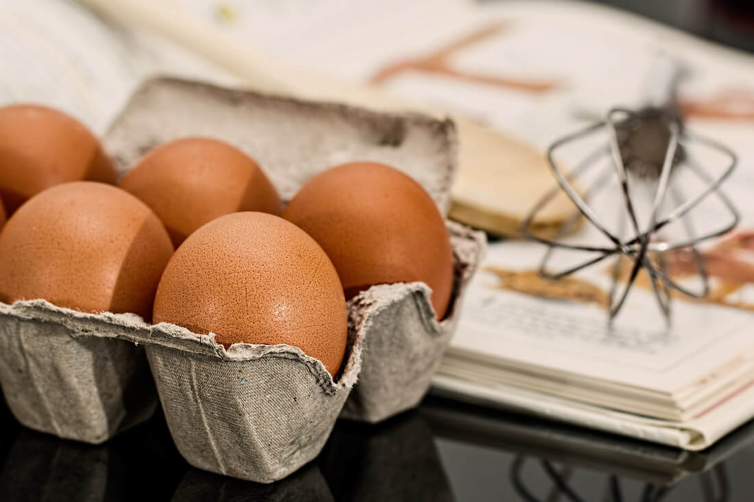 Eggs in a carton in front of a cookbook and utensils