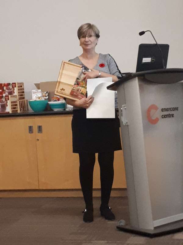 A woman holding a wooden cutting board and cookbook next to a podium