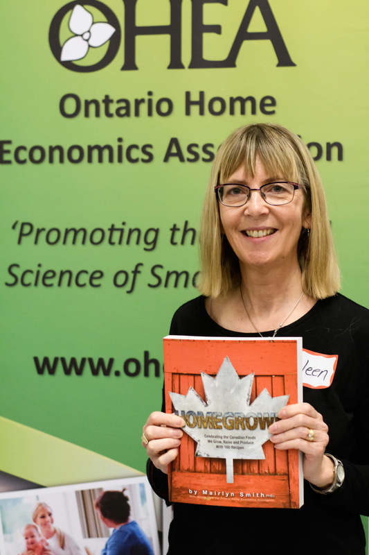 Eileen Stanbury, a Woman smiling holding a cookbook titled Homegrown with the OHEA banner behind her
